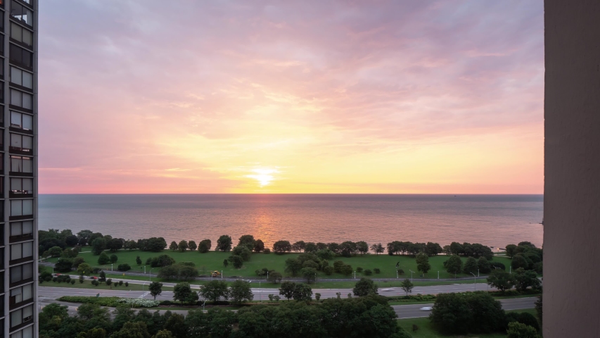 Incredible panning down aerial timelapse of a yellow pink purple and blue sunrise reflects on the water of Lake Michigan as it rises over the horizon with traffic zooming by on Lake Shore Drive below.