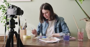 Artist, teenage girl, draws and records on video camera for his blog. Paints, drawings on a home table, camera on tripod, drawing and telling girl blogger vloger. Slider tracking shot