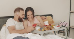 Happy young couple spending time and laughing in bed. Man and woman looking at smartphone screen and talking while having breakfast in early morning. Concept of leisure and relationship