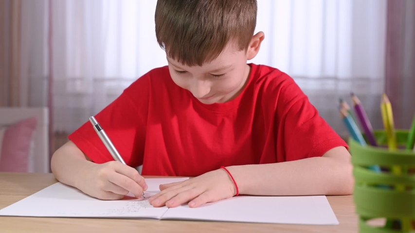 boy schoolchild with short haircut in red t-shirt writes in exercise book at table with pen holder against big window closeup