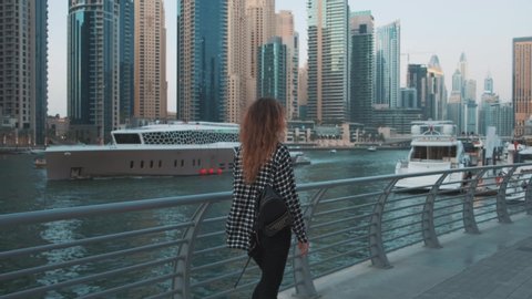 Young woman tourist walks promenade of Arab resort town. Redhead long flowing hair. Berth Dubai Marina modern white sea yachts. Girl enjoy beauty blue river water buildings skyscrapers. UAE 4k 2020