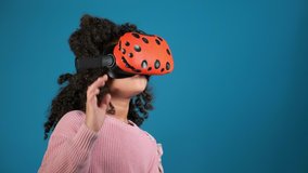 cute little African american girl in pink plays video game with virtual reality headset on blue background slow motion