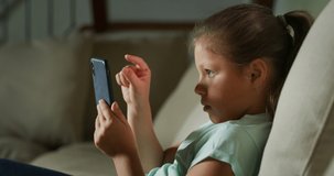 Authentic shot of a happy little girl is having fun to use a smartphone for playing games or study while sitting on a sofa in a living room at home.