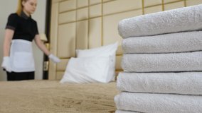 Hotel concept. Young, beautiful maid in uniform and apron making bed in room, shot through stack of fresh bath towels on bed sheet. Preparation of hotel room. 4 k video - Powered by Shutterstock - Get 15% off with code: PIKWIZARD15