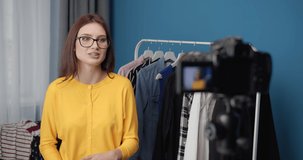 Smiling young lady in glasses and yellow blouse staying at home and talking on camera about trends. Popular blogger filming video for her social networks.