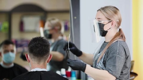  A hairdresser with security measures for Covid-19, cuts hair with a scissors to a man in a medicine mask, social distance, cutting hair with a medical mask, face shield , gloves