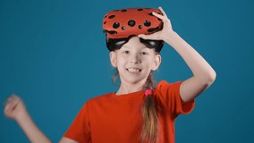 smiling little girl with blonde plait points by finger holding virtual reality glasses on head on blue background slow motion