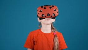 cute little girl in orange t-shirt with modern virtual reality goggles scared by VR scene on blue background slow motion