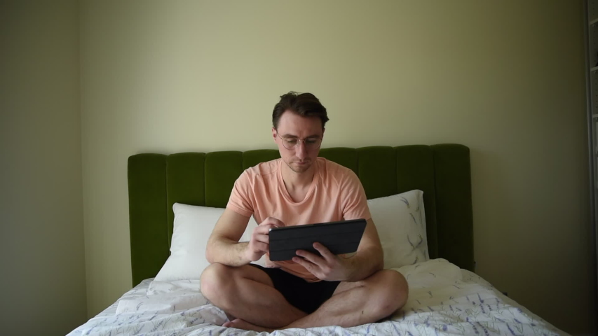 Young man sitting in bed and shopping online. Smiling beautiful man weared in pajama using digital tablet relax in bed, happy attractive man holding touchscreen tablet computer having fun surfing web.