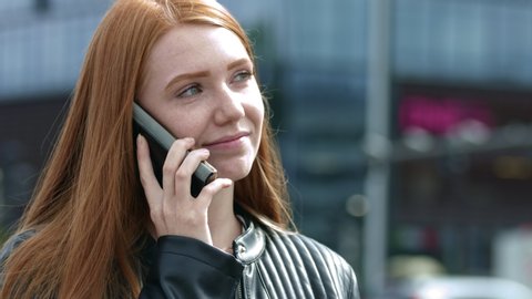 Happy and natural young ginger female talking over the phone in the public with a busy street in the background. Smiling and laughing while having a conversation.