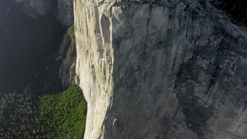 Vertical granite slopes of Mount El Capitan are in frame. Huge monolith, popular among rock climbers. one of the slopes is lit by the setting sun. Yosemite National Park. Aerial, 4K