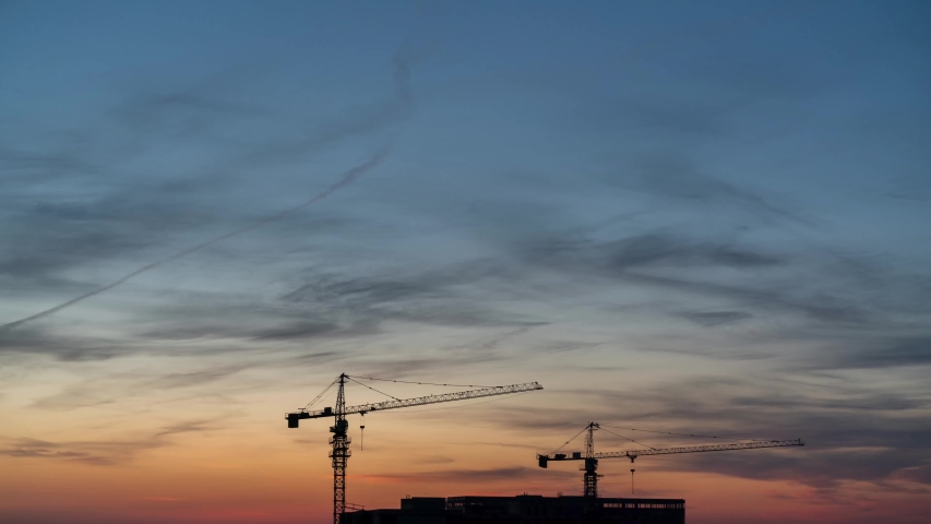 Time lapse of a beautiful orange and blue sunset with clouds on the background of the roof of a house under construction with cranes.