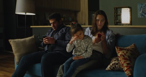 Parents stuck to their phones while sitting on a sofa with their daughter. Modern technology problem, social media addiction.