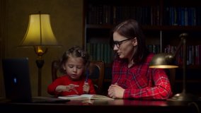 Preschool girl making homework with young 30s mother at home. Cute little girl writing down in notebook. Home education process. Mom and daughter in red dresses. 