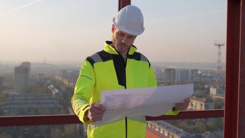 Male builder engineer man worker foreman architect in hard hat with drawing blueprint plan on construction site working at sunset. Industrial building city background outdoors. engineering 4 k Slow mo
