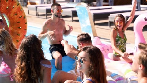 Friends have party in a private villa swimming pool. Happy young people in swimwear dancing, bonding and clubbing with floaties and inflatable mattress in luxury resort on sunny day. Tracking shot.