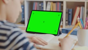 Smart Little Boy Uses Green Screen Chroma Key Laptop for Learning, Writes Down Useful Information. Distance Learning, e-Education, e-Learning, Homeschooling Concept. Close-up Over Shoulder POV 