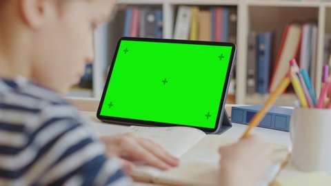 Smart Little Boy Uses Green Screen Chroma Key Laptop for Learning, Writes Down Useful Information. Distance Learning, e-Education, e-Learning, Homeschooling Concept. Close-up Over Shoulder POV 