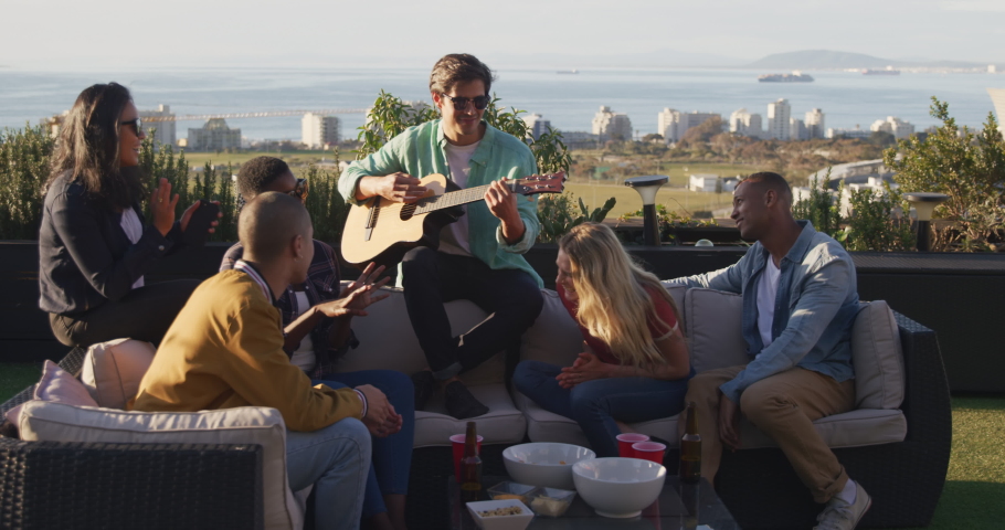 Multi-ethnic group of friends hanging out on a roof terrace together, Caucasian man is playing guitar, drinking drinks from red cups, drinking beer, in slow motion Royalty-Free Stock Footage #1052646467