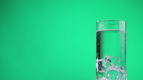 Glass full of cold sparkling water with ice cubes rotate on a green background. Transparent fizzy drink. Moving bubbles in soda. Refreshing mineral water.
