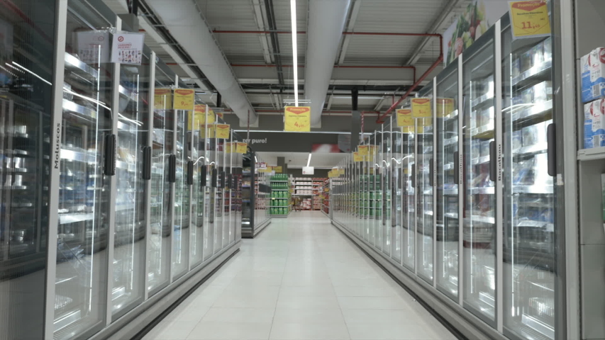 Frozen Food In Supermarket Aisle Tracking Shot. Tracking Shot of a big supermarket aisle with frozen food. People shopping in the background. Royalty-Free Stock Footage #1052659451