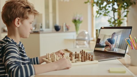 Brilliant Little Boy Playing Chess with His Chess Master, Uses Laptop for Video Call. Child Learns how To Play Chess Through Internet. Remote Online Education, E-Education. Over Shoulder