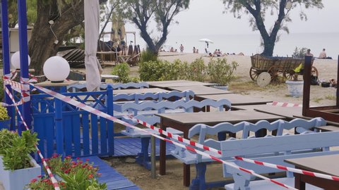 Neoi Epivates, Greece - May 16 2020: Empty Beach bars prepare to open for summer season. Closed outdoor bar restaurants with stacked chairs, tables & no access tape with bathers on sand by the sea.