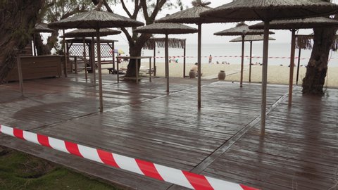 Neoi Epivates, Greece - May 16 2020: Empty Beach bars prepare to open for summer season. Closed outdoor bar restaurants with bamboo sun umbrellas, no access tape and bathers on sand by the sea.