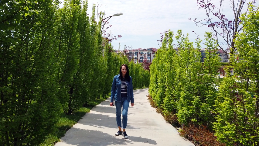 young woman with long dark hair in denim shirt and jeans walks along alley in city park. Sunny summer day. Gray asphalt road. Green alley of coniferous trees.
