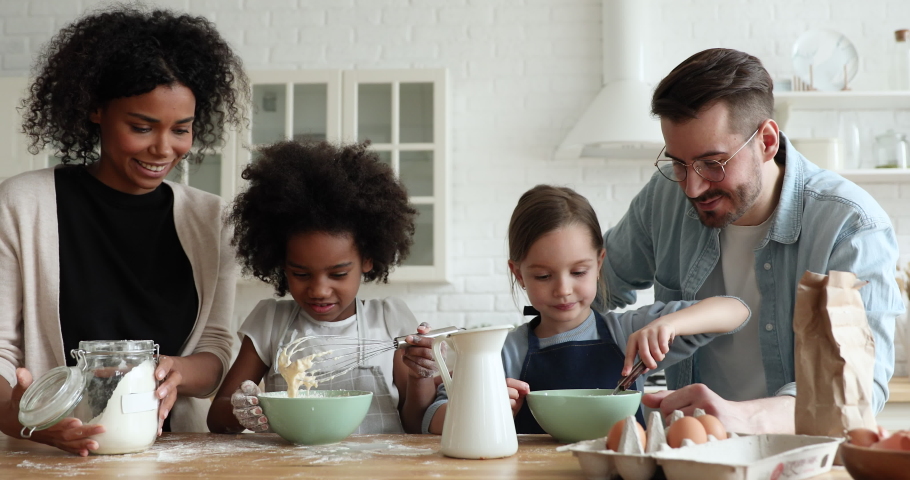 Cute mixed ethnicity little children daughters mixing flour, eggs and milk for holiday pancakes helping parents cooking together at home. Interracial family preparing homemade dough in kitchen. Royalty-Free Stock Footage #1052682233