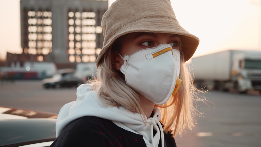 Portrait blonde caucasian girl wearing hat and protective face mask on parking space at sunset. Outbreak of coronavirus COVID-19. Pandemic. Health care. Royalty-Free Stock Footage #1052689445