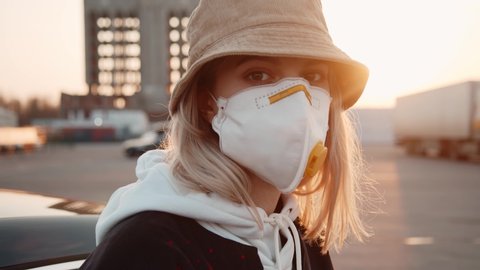 Portrait blonde caucasian girl wearing hat and protective face mask on parking space at sunset. Outbreak of coronavirus COVID-19. Pandemic. Health care.