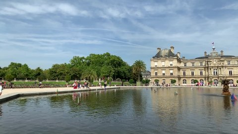 Footage of people walking and relaxing by pool at famous public garden called "Jardin Du Luxembourg" and palace in Paris. It is a sunny summer day. pans right.