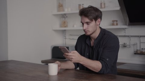 A young man sits at a table looking at his smartphone, drinking coffee, tea and smiling, the concept of spending time at home, leisure, isolation