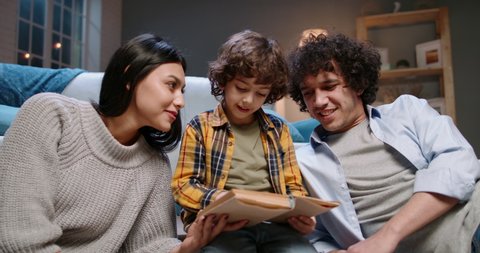 Cute young asian family spending time together at home. Little boy with curly hair reading a book with his parents, happily smiling - happy family, recreational pursuit concept 4k footage