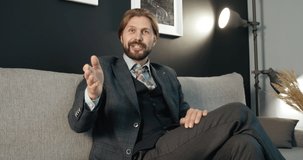 Confident mature businessman in trendy suit sitting on grey couch and talking on camera, first person view. Concept of experience and business idea