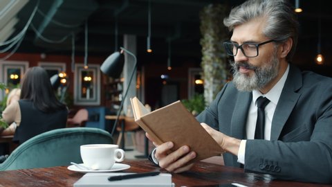 Mature man in glasses and suit is reading book turning pages at table in restaurant focused on literature. Leisure time activity and smart people concept.