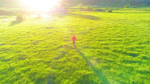 UHD 4K aerial view. Low altitude flight near sporty man running at perfect green grass rural meadow offroad. Sunrise in mountain. Green farm trial path and sun rays on horizon. Fast horizontal