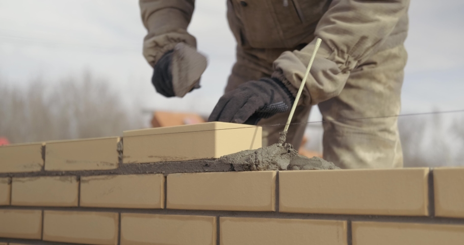 Bricklayer works at construction site, lays bricks in masonry, builds house, erects brick wall, yellow cladding brick, works as trowel, open air, in special uniform, professional work, smooth seams | Shutterstock HD Video #1052709719