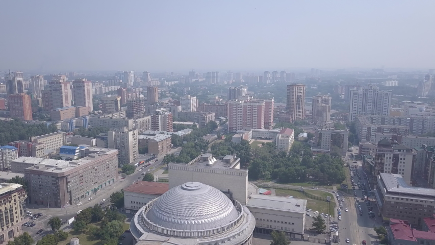 Novosibirsk State Academic Theater of Opera and Ballet. A monumental circular building crowned by a massive silver-toned dome dominates the center of a broad urban square, surrounded by a grid of mid-rise residential and office towers under a pale summer sky. The theater