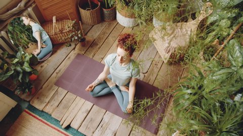 Top view of young redhead woman in sportswear sitting in lotus pose on yoga mat and holding hands in mudras while meditating at home in room filled with green plants