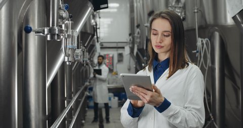 Crop view of good looking female worker touching display while entering data in tablet. Woman and man in white lab coat checking equipment gauges and standing at rows of brewing vats.
