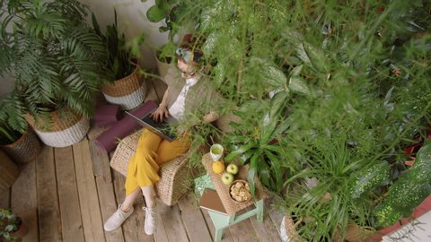Top view shot of young woman sitting on rattan chair and typing on laptop while working remotely from home in cozy room with lots of green plants