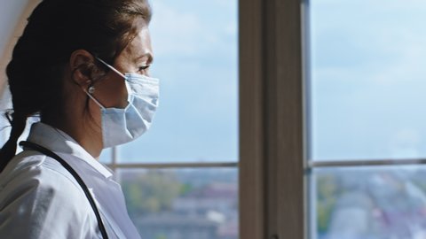 Portrait of attractive woman doctor with a protective mask looking through the window and feeling very tired