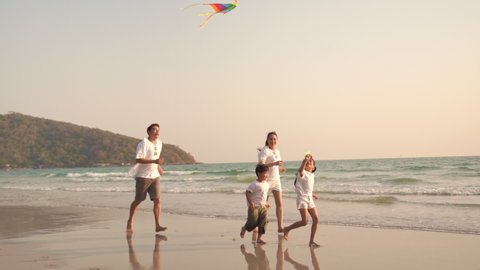 Slow motion of smiling Asian family parent with two child boy and girl running and playing kite together on the beach at summer sunset. Happy family relax and having fun in holiday vacation trip