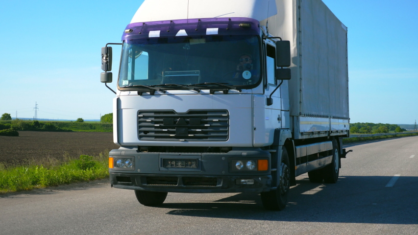 Camera follows to truck with cargo trailer driving on highway and transporting goods at summer day. Lorry riding through countryside with nature landscape at background. Slow mo Front view Close up | Shutterstock HD Video #1052729696
