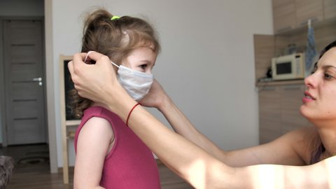 Mom tries on a medical mask for her little daughter. Stay home mom. Coronavirus or COVID-19. Cute little girl in a protective mask at home. Mom teaches daughter to put on a medical mask