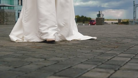 CU, slow motion: graceful bride in long white silk wedding dress walks to groom along spacious city square on summer day close low angle shot