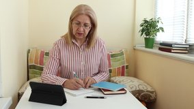 Mature woman study over the Internet, writes tasks and looks in a digital tablet. Online education for adults.