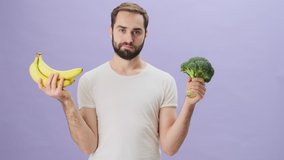 A positive attractive young man wearing a white t-shirt is choosing between banana and broccoli and give a broccoli to the viewer standing isolated over gray background